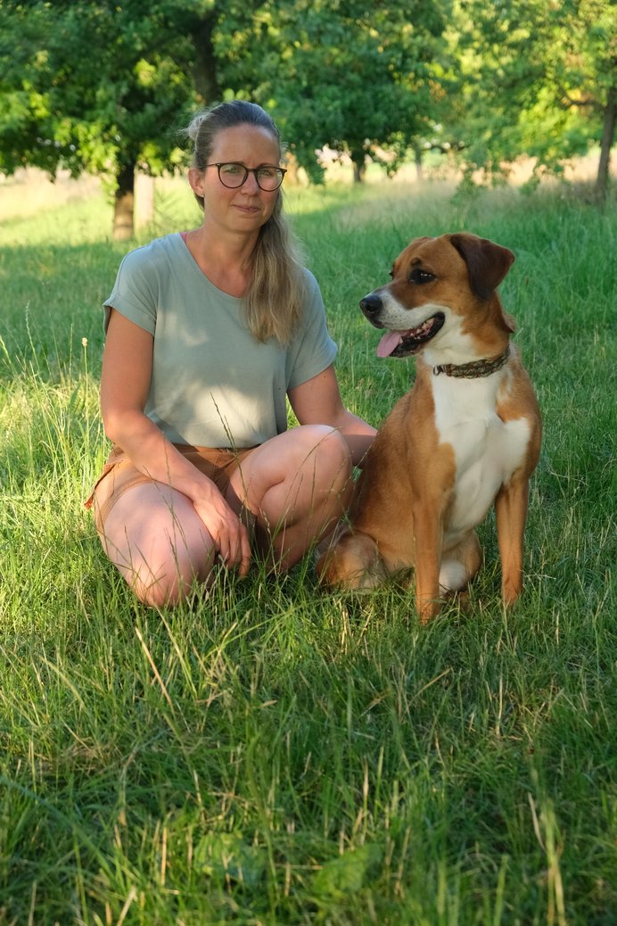 Frau mit Brille in T-Shirt und kurzer Hose mit braun-weißem kurzhaarigen Hund neben sich auf einer Wiese kniend.