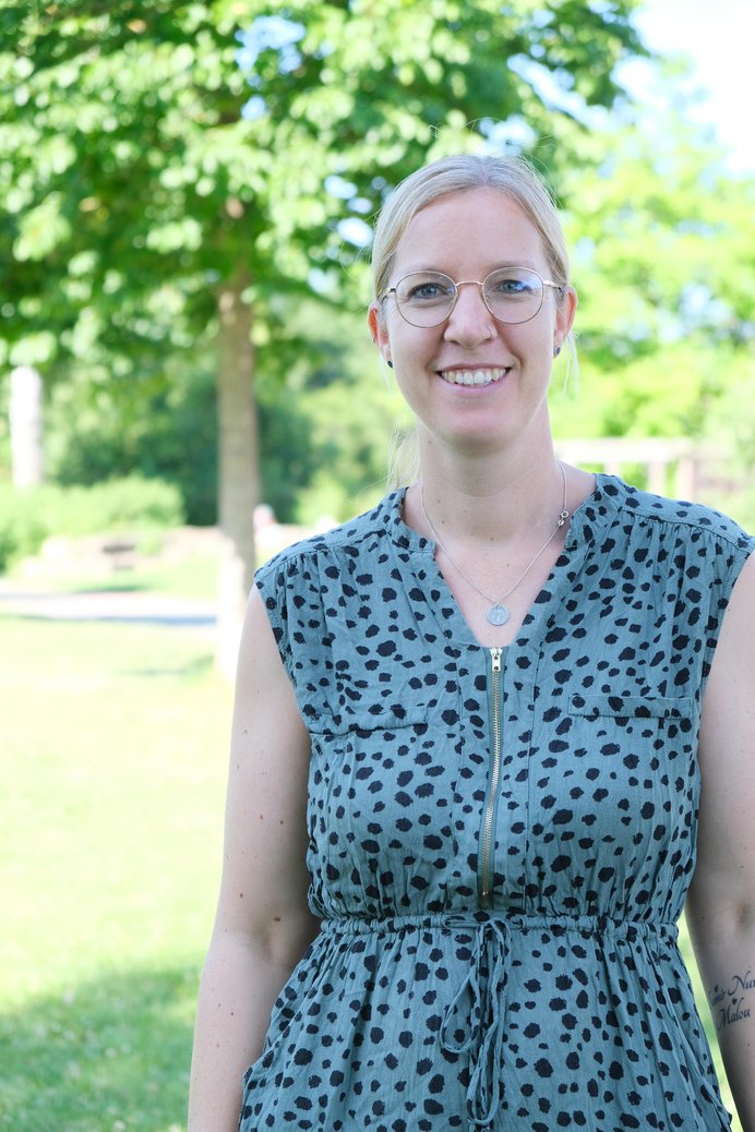 Frau mit blonden zusammengebundenen Haaren, Brille und Kleid im Freien.