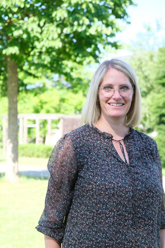 Nahaufnahme einer Frau mit blonden halblangen haaren und Brille im Freien mit Baum und Büschen im Hintergrund. 